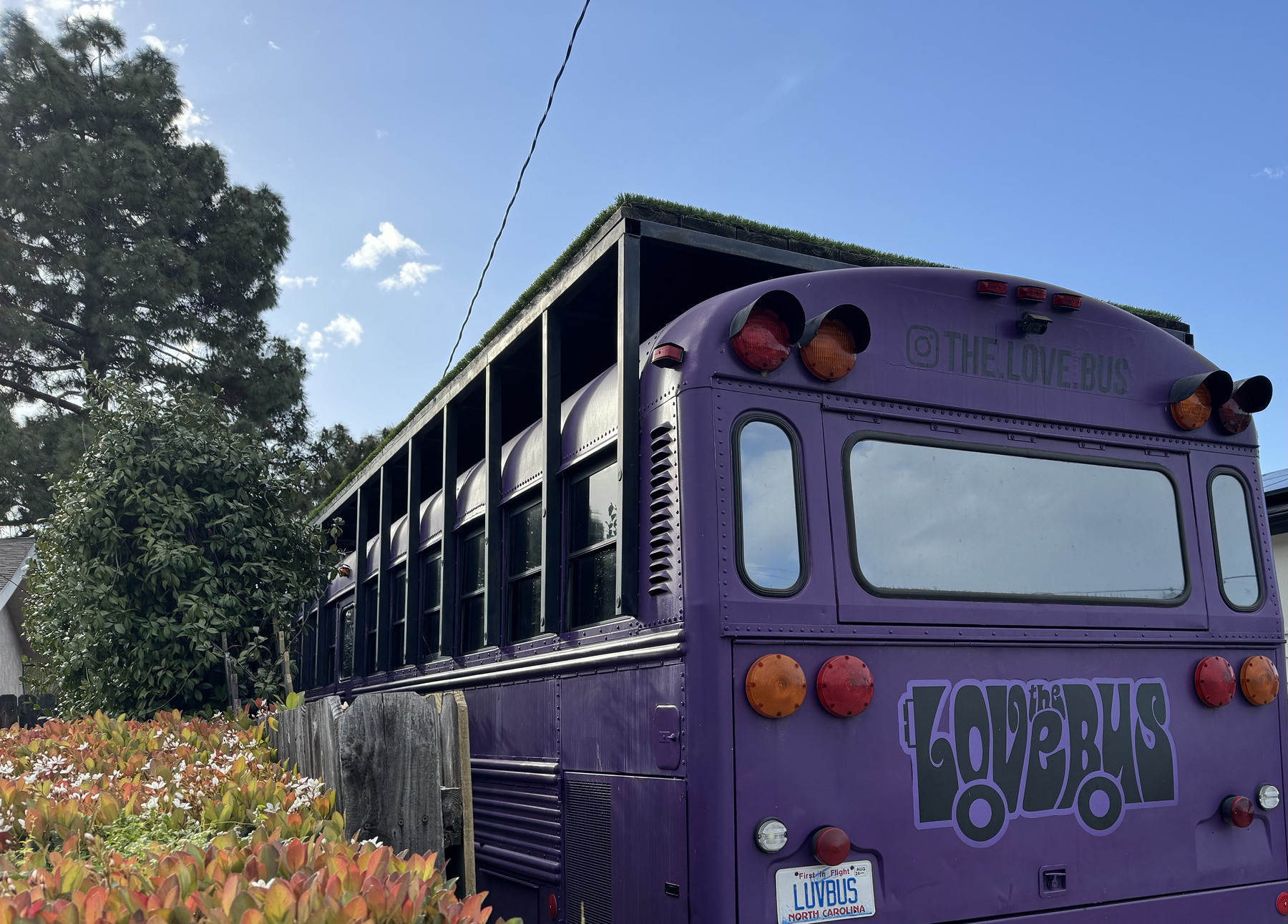 School bus painted deep purple with a platform on top and the logo "The Love Bus" in a groovy 1960s font on the rear door. 