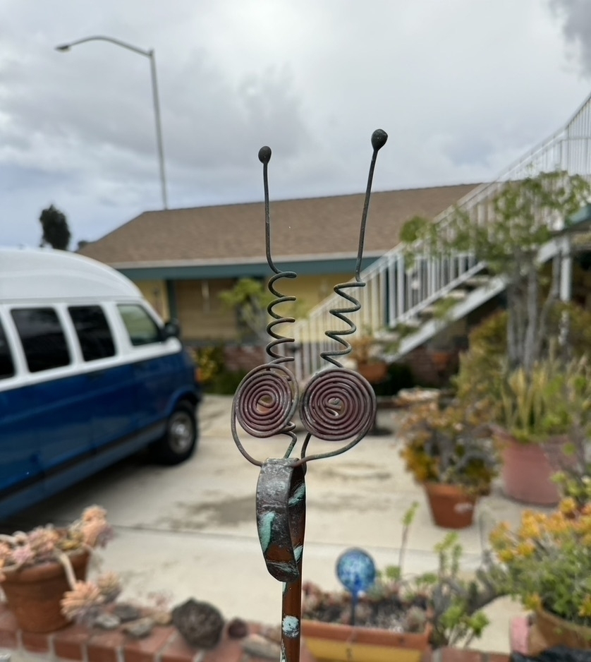 Artsy photo of the front yard of a suburban house. Dominating the foreground is a cute wire sculpture of an insect face. In the background, a blue-and-white minivan and house, slightly blurry. 