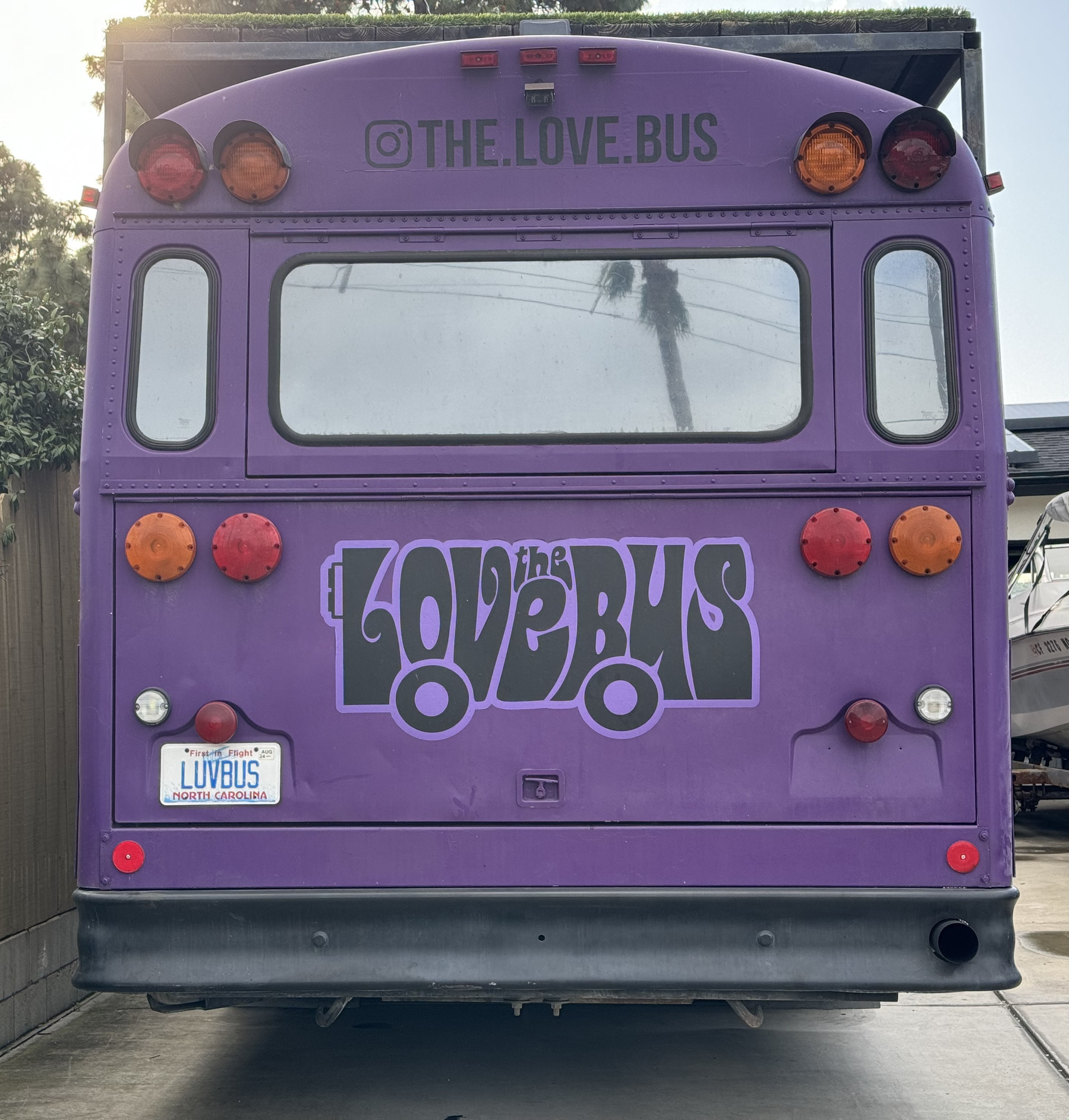 Alternate angle of the same school bus, painted deep purple with a platform on top and the logo "The Love Bus" in a groovy 1960s font on the rear door.